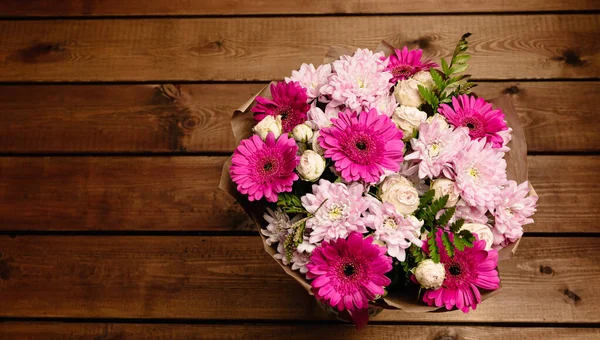 A large beautiful bouquet of chrysanthemums, gerberas, roses and ferns in pink and purple colors wrapped in brown craft paper on a wooden background. Postcard for the holiday. Close-up. Banner.