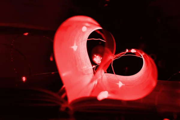 Red art book novel with sheets in the shape of a heart decorated with bright garland lights and highlights in the background, a romantic symbol of love, macro. Valentine day. Greeting card. Close up.