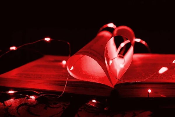 Red art book novel with sheets in the shape of a heart decorated with bright garland lights and highlights in the background, a romantic symbol of love, macro. Valentine day. Greeting card. Close up.