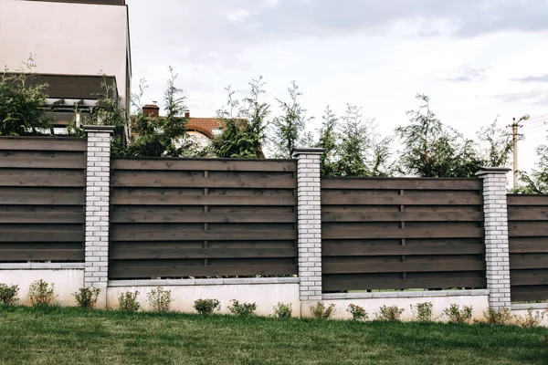 Horizontal tiered sections of brown wooden boards fence and white brick pillars. Live plantings. Green thuja bushes and lawn. Territory landscaping. Capital fencing. Hedge and wall on a country house.