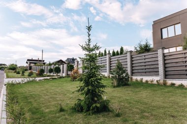 Horizontal sections of brown wooden boards fence and white brick pillars. Live plantings. Green spruce, pine, bushes and lawn. Territory landscape design. Capital fencing. Landscaping and gardening.
