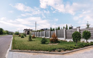 Horizontal tiered sections of brown wooden boards fence and white brick pillars. Live plantings. Green thuja, pine, bushes and lawn. Territory landscaping. Capital fencing. Street. Landscape design.