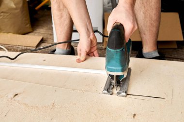 İnşaat işçisinin erkek elleri kontraplaktan elektrikli testereyle parçalar kesip biçiyor. Kesme işlemi. İç dekorasyonun demir testeresiyle yapıldığı bina. Kiralık araç. Uzayı kopyala Ekipman.