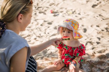 Güzel, genç bir anne ve iki yaşındaki bir kız çocuğu kumların üzerinde oturup karpuz yiyorlar. Sahilde piknik. Deniz yolculuğu tatili. Aile bakımı. Mutluluk yaşam tarzı. Sevgi. Yakın plan portre.