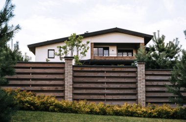 Yeşil bitkilerle çevrili ahşap bir çitin parçası. Evergreen çiti. Yatay çizgiler. Tuğla. İskandinav tarzı arka bahçe. Batı sınırı. Ev güvenliği bariyeri. Peyzaj tasarımı. Düzeltilmemiş tahta.