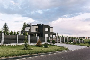 Geniş panoramik pencereli, küp şeklinde modern iki katlı kır evi. Tahta çitler ve tuğla direkler. Giriş grubu. Yeşillik bahçeli, yeşil çimenlikli. Kırsal kesimde. Peyzaj tasarımı.