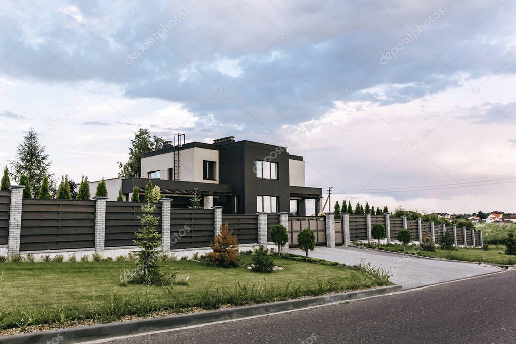 Modern two-storey country house in cube shape with large panoramic windows. Wooden fence and brick posts. Entry group. Front and back yard with evergreens, green lawn. Countryside. Landscape design.