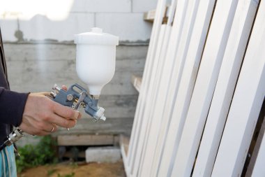 Bir adamın elindeki sprey tabancaya yakın çekim. Tahtaları beyaz boyayla boyama işlemi. Bitiriyorum. Eski mobilyaların restorasyonu. Kompresör operasyonu. İş aleti. Hızlı ve kolay. Boşluğu kopyala.