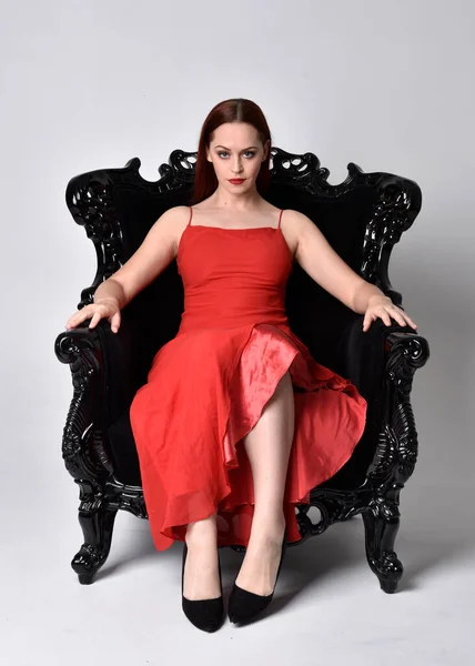 赤いセクシーなドレスを着た可愛い女性の完全な長さの肖像画 黒の華やかなアームチェアに腰掛け スタジオを背景に — ストック写真