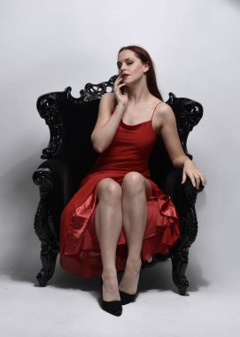 Kırmızı seksi elbise giyen güzel bir kadının uzun bir portresi. Stüdyonun arka planında, siyah bir koltukta otururken..