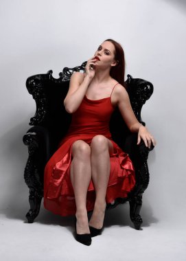 Kırmızı seksi elbise giyen güzel bir kadının uzun bir portresi. Stüdyonun arka planında, siyah bir koltukta otururken..