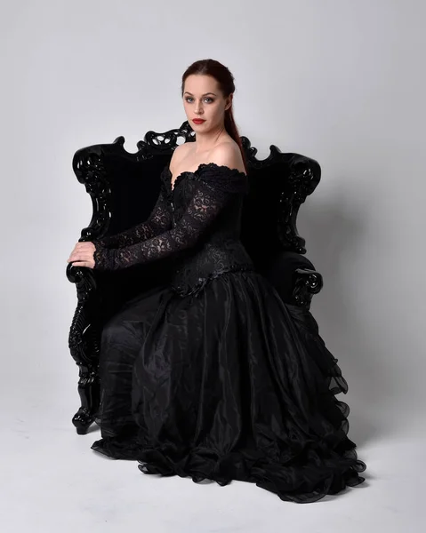 黒いゴシックのドレスを着た女性の完全な長さの肖像画は 華やかな黒いアームチェアに座っています 座席のポーズ スタジオの背景を背景に — ストック写真