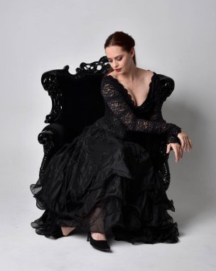 Siyahi gotik elbiseler giyen, süslü siyah bir koltukta oturan bir kadının tam boy portresi. Oturmuş poz, stüdyo arka planında..