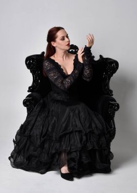 Siyahi gotik elbiseler giyen, süslü siyah bir koltukta oturan bir kadının tam boy portresi. Oturmuş poz, stüdyo arka planında..