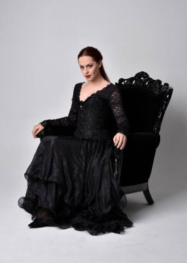 Siyahi gotik elbiseler giyen, süslü siyah bir koltukta oturan bir kadının tam boy portresi. Oturmuş poz, stüdyo arka planında..