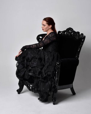 Siyahi gotik elbiseler giyen, süslü siyah bir koltukta oturan bir kadının tam boy portresi. Oturmuş poz, stüdyo arka planında..