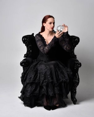 Siyahi gotik elbiseler giyen, süslü siyah bir koltukta oturan bir kadının tam boy portresi. Oturmuş poz, stüdyo arka planında..