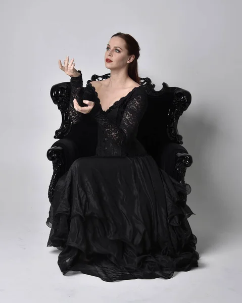 黒いゴシックのドレスを着た女性の完全な長さの肖像画は 華やかな黒いアームチェアに座っています 座席のポーズ スタジオの背景を背景に — ストック写真
