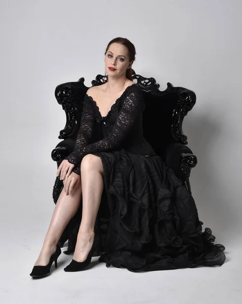 穿着黑色哥特式连衣裙的女人的全长肖像 坐在华丽的黑色扶手椅上 在工作室背景下的坐姿 — 图库照片