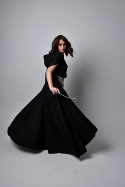 Uzun boylu, esmer, uzun siyah pelerinli, etekli ve çizmeli güzel bir kadın portresi. Stüdyonun arka planında sihirli bir değnek tutuyor..