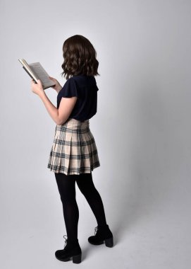 Uzun boylu, esmer, etek ve çizme giyen güzel bir kadın portresi. Stüdyo arkaplanına karşı kitap tutarak poz vermek.