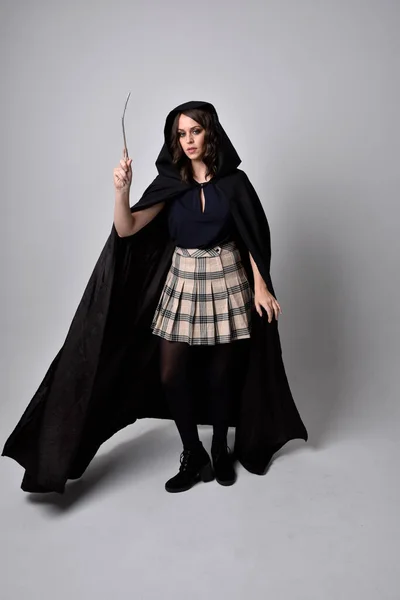 タータンスカートと長い黒のマントを着た美しいブルネットの女性の完全な長さの肖像画 魔法の杖をスタジオの背景に持って立ちポーズ — ストック写真