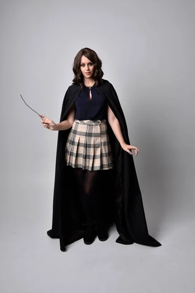 タータンスカートと長い黒のマントを着た美しいブルネットの女性の完全な長さの肖像画 魔法の杖をスタジオの背景に持って立ちポーズ — ストック写真