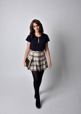 Uzun boylu, esmer, etek ve çizme giyen güzel bir kadın portresi. Stüdyo arkaplanına karşı kitap tutarak poz vermek.