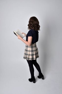 Uzun boylu, esmer, etek ve çizme giyen güzel bir kadın portresi. Stüdyo arkaplanına karşı kitap tutarak poz vermek.