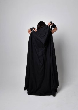 Uzun siyah saçlı, uzun siyah cüppeli güzel bir kadın portresi. Stüdyonun arka planında kameraya dönük poz vermek.