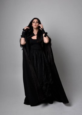 Uzun siyah saçlı, uzun siyah cüppeli güzel bir kadın portresi. Stüdyonun arka planında kameraya dönük poz vermek.