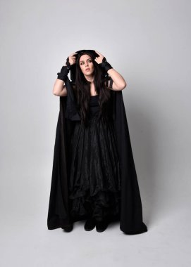 Uzun siyah saçlı, uzun siyah cüppeli güzel bir kadın portresi. Stüdyonun arka planında kameraya dönük poz vermek.