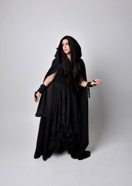 Uzun siyah saçlı, uzun siyah cüppeli güzel bir kadın portresi. Stüdyonun arka planında kameraya dönük poz vermek.