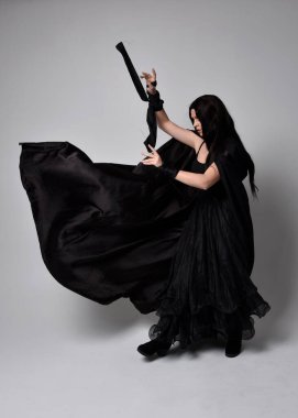 Uzun siyah saçlı, uzun siyah cüppeli güzel bir kadın portresi. Stüdyonun arka planında kameraya dönük poz vermek.