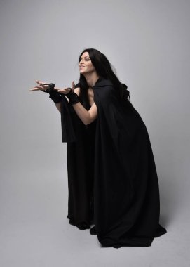 Uzun siyah saçlı, uzun siyah cüppeli güzel bir kadın portresi. Stüdyonun arka planında kameraya dönük poz vermek.