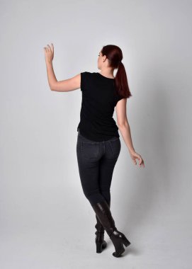 Kırmızı saçlı, at kuyruklu, siyah tişörtlü ve kot pantolonlu bir kadının tam boy portresi. Ayakta durarak, eller kameraya uzanarak stüdyonun arka planına doğru..
