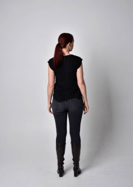 Sade, uzun, at kuyruklu, siyah tişört ve kot pantolon giyen kızıl saçlı bir kadın portresi. Kameraya dönük poz vermek, kamera, stüdyonun arka planında..