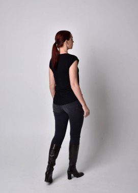 Sade, uzun, at kuyruklu, siyah tişört ve kot pantolon giyen kızıl saçlı bir kadın portresi. Kameraya dönük poz vermek, kamera, stüdyonun arka planında..