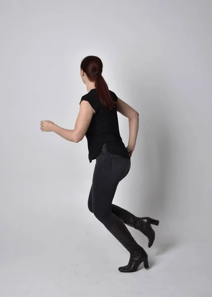 Simple full length portrait of woman with red hair in a ponytail, wearing casual black tshirt and jeans. Standing pose with back to the camera the camera, against a  studio background.