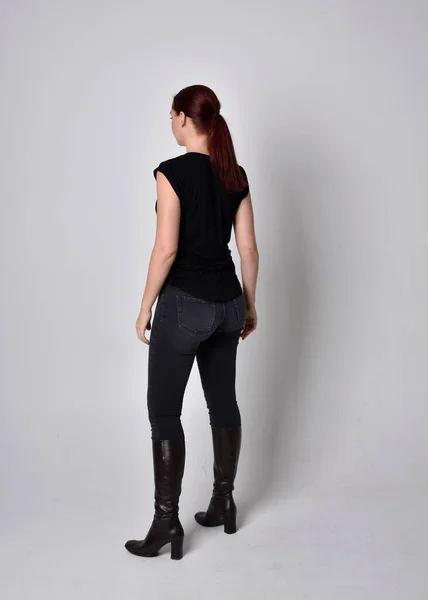 Simple full length portrait of woman with red hair in a ponytail, wearing casual black tshirt and jeans. Standing pose with back to the camera the camera, against a  studio background.