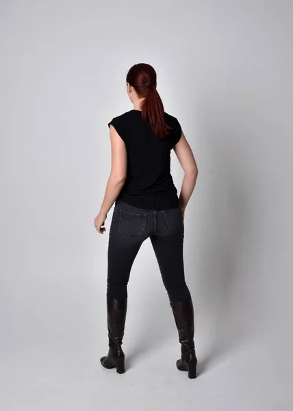 Simple full length portrait of woman with red hair in a ponytail, wearing casual black tshirt and jeans. Standing pose with back to the camera the camera, against a  studio background.