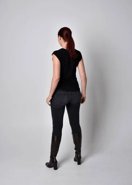 ポニーテールに赤い髪の女性のシンプルな全長の肖像画 カジュアルな黒のTシャツとジーンズを身に着けています カメラに背を向けて立ちポーズ スタジオの背景を背景に — ストック写真