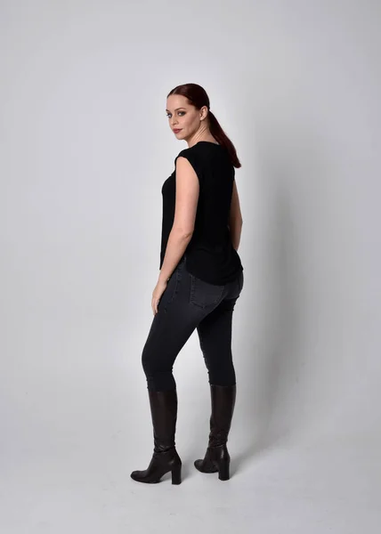 ポニーテールに赤い髪の女性のシンプルな全長の肖像画 カジュアルな黒のTシャツとジーンズを身に着けています カメラに背を向けて立ちポーズ スタジオの背景を背景に — ストック写真