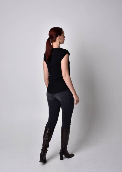 Simple full length portrait of woman with red hair in a ponytail, wearing casual black tshirt and jeans. Standing pose with back to the camera the camera, against a  studio background.