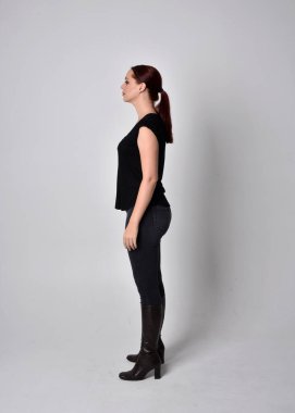 Sade, uzun, at kuyruklu, siyah tişört ve kot pantolon giyen kızıl saçlı bir kadın portresi. Stüdyonun arka planında yan profil ile poz vermek.
