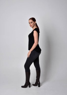 Sade, uzun, at kuyruklu, siyah tişört ve kot pantolon giyen kızıl saçlı bir kadın portresi. Stüdyonun arka planında yan profil ile poz vermek.