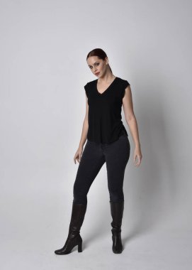 Sade, uzun, at kuyruklu, siyah tişört ve kot pantolon giyen kızıl saçlı bir kadın portresi. Stüdyonun arka planında el hareketleriyle poz vermek.