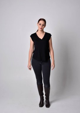 Sade, uzun, at kuyruklu, siyah tişört ve kot pantolon giyen kızıl saçlı bir kadın portresi. Stüdyonun arka planında öne doğru poz veriyor..