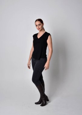 Sade, uzun, at kuyruklu, siyah tişört ve kot pantolon giyen kızıl saçlı bir kadın portresi. Stüdyonun arka planında öne doğru poz veriyor..