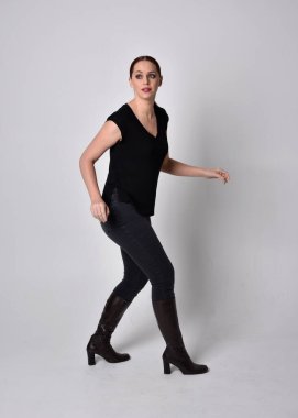 Sade, uzun, at kuyruklu, siyah tişört ve kot pantolon giyen kızıl saçlı bir kadın portresi. Stüdyonun arka planında öne doğru poz veriyor..